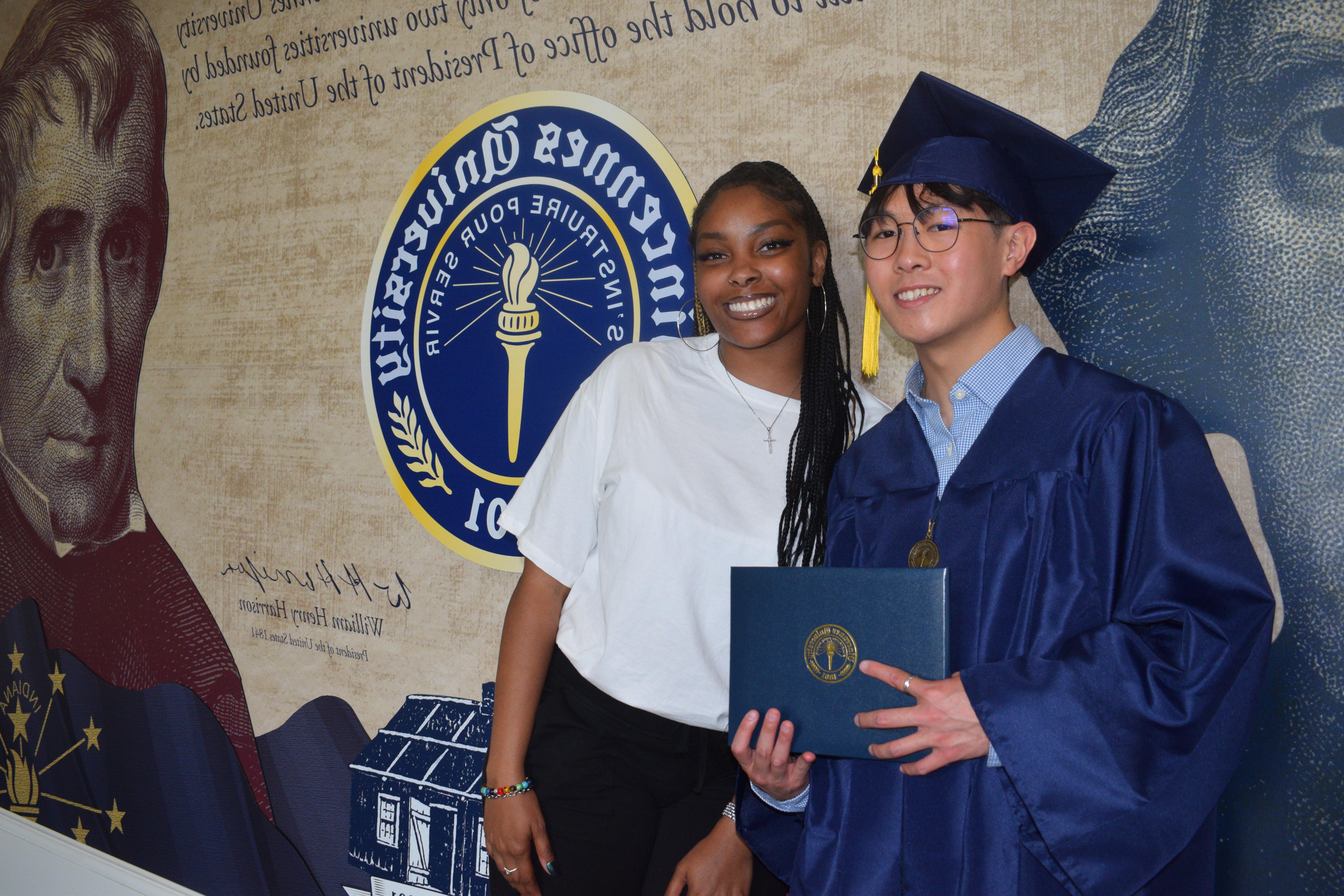 撒母耳 Lo wears a graduation cap and gown while holding a 澳门足球博彩官方网址 degree cover. A female student stands next to him. The students stand in front of a 澳门足球博彩官方网址 mural featuring Thomas Jefferson, 威廉·亨利·哈里森, and the 澳门足球博彩官方网址 Seal plus Indiana and American flags.